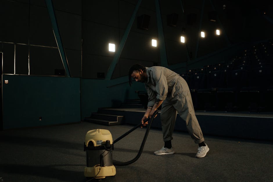 A person wearing a grey jumpsuit is operating a large vacuum cleaner on a dark, empty auditorium or theatre stage with black flooring and tiered seating in the background. The cleaning process appears